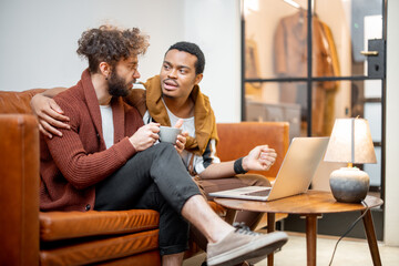 Gay couple with different nationality having close conversation while sitting on a couch at home. Concept of homosexual relations and lifestyle at home. Caucasian and hispanic man together indoors