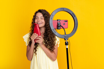 Teen girl hold hair conditioner or shampoo bottle in front of camera for vlog isolated on yellow background. Teenager working as blogger, recording video blog.