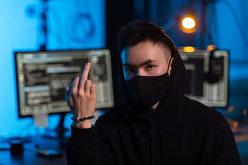 Poster - cybercrime, hacking and technology concept - hacker wearing black mask and hood showing middle finger in dark room with computers at night