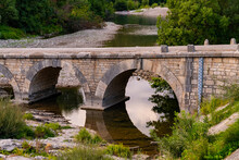 Old Bridge Spanning Over Cèze River In Provence South France Near Idyllic Village Montclus. Evening Atmosphere With Brick Arches Reflected On Water Surface. Very Low Water Level Caused By Drought.