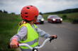Rear view of little girl in reflective vest riding bike on road with cars behind her, road safety education concept.