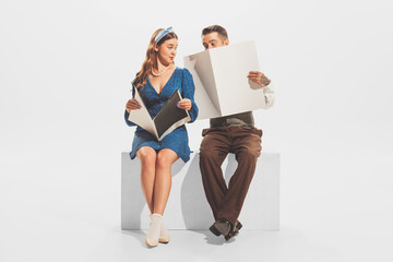 Portrait of young man and woman in retro outfit sitting together, reading magazine or newspaper isolated over white studio background