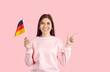 © Studio Romantic - Happy cheerful beautiful young German woman in casual sweatshirt standing isolated on pastel pink background, holding flag of Germany, pointing her index finger at copy space on the right, and smiling