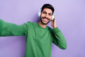 Sticker - Photo of toothy beaming cheerful man wear green pullover doing selfie listen music in headphones isolated on violet color background