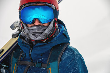 Equipped skier holding skis on his shoulder and looking directly at the camera, portrait. Man with winter equipment in the mountains at a ski resort