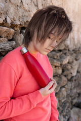 Wall Mural - Short-haired Teenage girl in pink hoodie staying near a stone wall with red water bottle