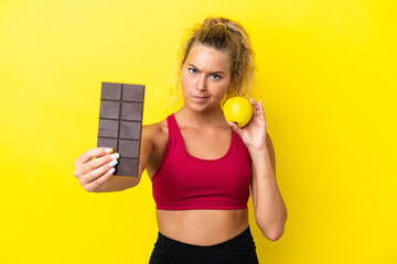 Wall Mural - Girl with curly hair isolated on yellow background taking a chocolate tablet in one hand and an apple in the other