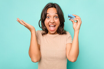 Wall Mural - Young hispanic woman holding stapler isolated on blue background receiving a pleasant surprise, excited and raising hands.