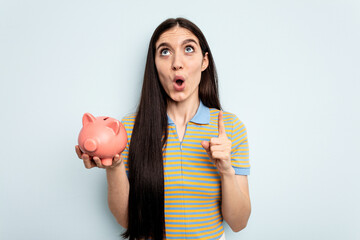 Young caucasian woman holding a piggy bank isolated on blue background pointing upside with opened mouth.