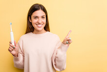 Young Caucasian Woman Holding A Electric Toothbrush Isolated On Yellow Background Smiling And Pointing Aside, Showing Something At Blank Space.