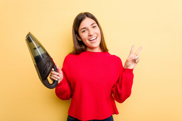 Wall Mural - Young caucasian woman holding a hand vacuum cleaner isolated on yellow background joyful and carefree showing a peace symbol with fingers.