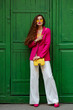 Fashionable woman wearing trendy outfit with yellow sunglasses, bag, pink blazer, wide leg white trousers, posing near green door. Full-length outdoor portrait
