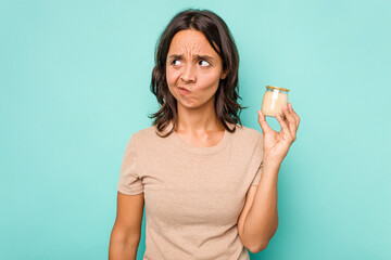 Wall Mural - Young hispanic woman holding yogurt isolated on blue background confused, feels doubtful and unsure.