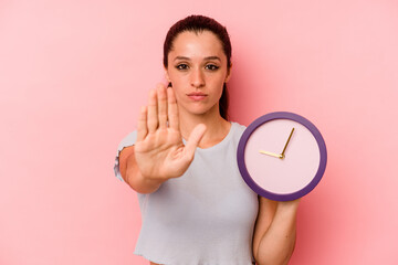 Wall Mural - Young caucasian woman holding a clock isolated on pink background standing with outstretched hand showing stop sign, preventing you.