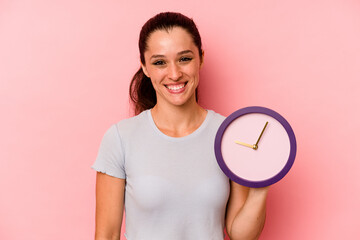 Wall Mural - Young caucasian woman holding a clock isolated on pink background happy, smiling and cheerful.