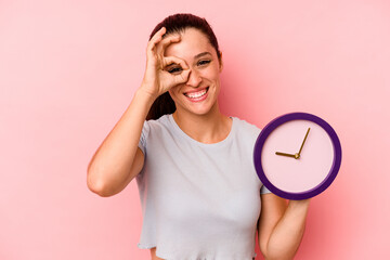 Wall Mural - Young caucasian woman holding a clock isolated on pink background excited keeping ok gesture on eye.