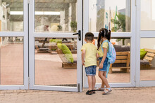 Little Boy And Girl With A Tie-dye Color Backpack Looking To The Corridor Of The School. Little Kids Peeping Through The Glass Door.