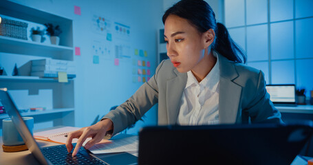 Wall Mural - Young Asia cheerful professional woman sitting on desk focus concentrate working on laptop and digital tablet check detail final work in office at night. Lady business suit, Startup business concept.