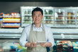© Liubomir - portrait of handsome asian staff man salesman in apron standing and looking at camera in grocery store supermarket. Male worker seller or small business owner. indoor. food, groceries market smile