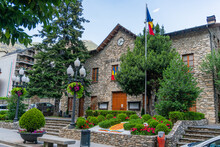 Sant Julia De Loria Town Hall In Andorra, A Stone Building With The Andorran Flag On A Mast And A Garden With Trees And Flowers In Summer, With The Mountains Behind And Closed Wooden Doors