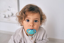 Curious Little Boy With Blue Pacifier Looking At Camera With Interest And Sitting On Floor In Living Room At Home