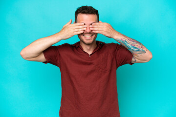 Young caucasian handsome man isolated on blue background covering eyes by hands and smiling