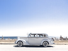 Vintage Old Car Along The Beach In Malibu.  Pacific Ocean In The Background.  Balancing Rocks To The Side.  White Packard Vehicle.