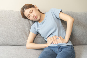 Wall Mural - Flatulence asian young woman hand in stomach ache, suffer from food poisoning, abdominal pain and colon problem, gastritis or diarrhoea lying on sofa at home. Patient belly, abdomen or inflammation.