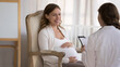 © fizkes - Young smiling pregnant woman patient answers questions of gynecologist during visit at home. Ob-gyn asks about health in late stages of pregnancy, filling medical form, provide prenatal care concept
