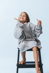 Playful cheerful little girl in oversize hoodie sitting on a stepping stool. Against bluish white background