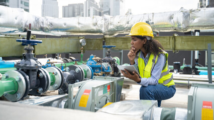 Service engineer woman dark skin wearing uniform and safety helmet under inspection and checking production process,HVAC system (Heating,Ventilation and Air Conditioning) on factory station by tablet.