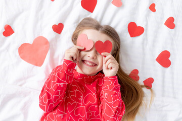 the concept of Valentine's day, a sweet child girl lying on a white bed at home in red pajamas among hearts covers her face with hearts, indulges and fools around, smiles with happiness