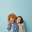 © wayhome.studio  - Vertical shot of mixed race women point fingers above at copy space show place for your text or design laugh positively share cool link focused overhead isolated over blue background. Look there