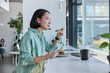 © wayhome.studio  - Young ethic woman enjoys eating tasty lunch poses in cozy cafeteria at windowsill surrounded by modern technologies for working freelance wears spectacles and casual shirt looks thoughtfully forward