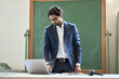 © insta_photos - Busy young indian business man working on laptop standing in office at work. Arab teacher professor coach thinking on online class mentoring using computer technology for remote teaching in classroom