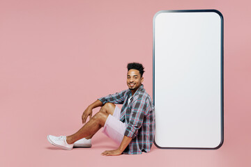 Wall Mural - Full body young man of African American ethnicity in blue shirt sit big huge blank screen mobile cell phone copy space mockup area workspace look camera isolated on plain pastel light pink background.