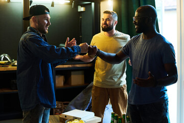 Three young cheerful buddies in casualwear making punch bump over table with bottles of beer and snacks while gathering in garage