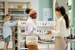 © pressmaster - Young black woman asking shop assistant for advice of cosmetic products while both standing in front of display with makeup goods