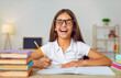 © Studio Romantic - Happy, positive child having fun while doing school homework. Portrait of a cheerful student girl in glasses laughing while writing in her notebook at her desk at home. Education and learning concept