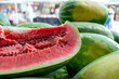 Fresh elongated watermelon with a transverse cut showing a pink chunky inside with lots of black seeds and striped green and yellow exterior rind with lots of whole melons on a table for sale.   