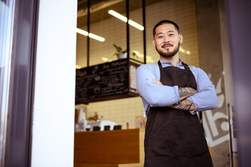 Positive asian owner in apron crossed arms near entrance of cafe 