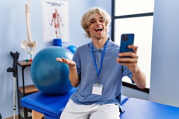 Sticker - Young man working at rehabilitation clinic doing video call with smartphone celebrating achievement with happy smile and winner expression with raised hand