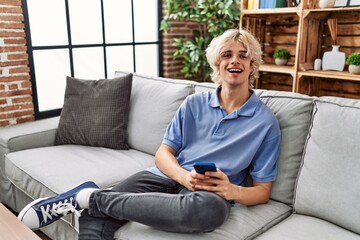 Poster - Young blond man using smartphone sitting on sofa at home