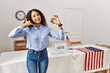 Beautiful hispanic woman standing by at political campaign by voting ballot smiling doing phone gesture with hand and fingers like talking on the telephone. communicating concepts.