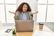 Beautiful hispanic business woman sitting on desk at office working with laptop clueless and confused expression with arms and hands raised. doubt concept.