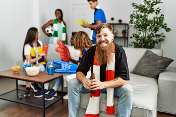 Wall Mural - Young soccer hooligan man smiling happy drinking beer at home.