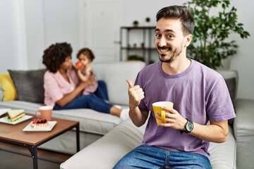 Wall Mural - Hispanic father of interracial family drinking a cup coffee pointing to the back behind with hand and thumbs up, smiling confident