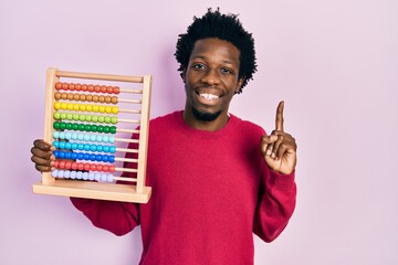 Wall Mural - Young african american man holding traditional abacus smiling with an idea or question pointing finger with happy face, number one