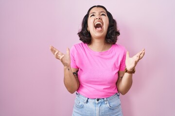 Poster - Young hispanic woman standing over pink background crazy and mad shouting and yelling with aggressive expression and arms raised. frustration concept.