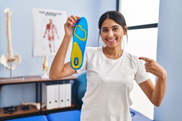 Wall Mural - Young hispanic woman holding shoe insole at physiotherapy clinic smiling happy pointing with hand and finger